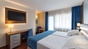 A hotel room with a TV mounted on the wall, a desk with drawers, two beds with white linens, a blue chair, a small table, and a window with blue curtains.