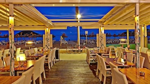 An outdoor restaurant with wooden tables and wicker chairs, overlooking a beach with umbrellas and a sunset view. - Flamboyan Caribe Hotel & Spa (Calvia)