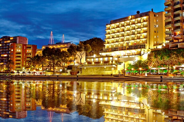 A waterfront hotel with multiple floors, balconies, and a reflection in the water.