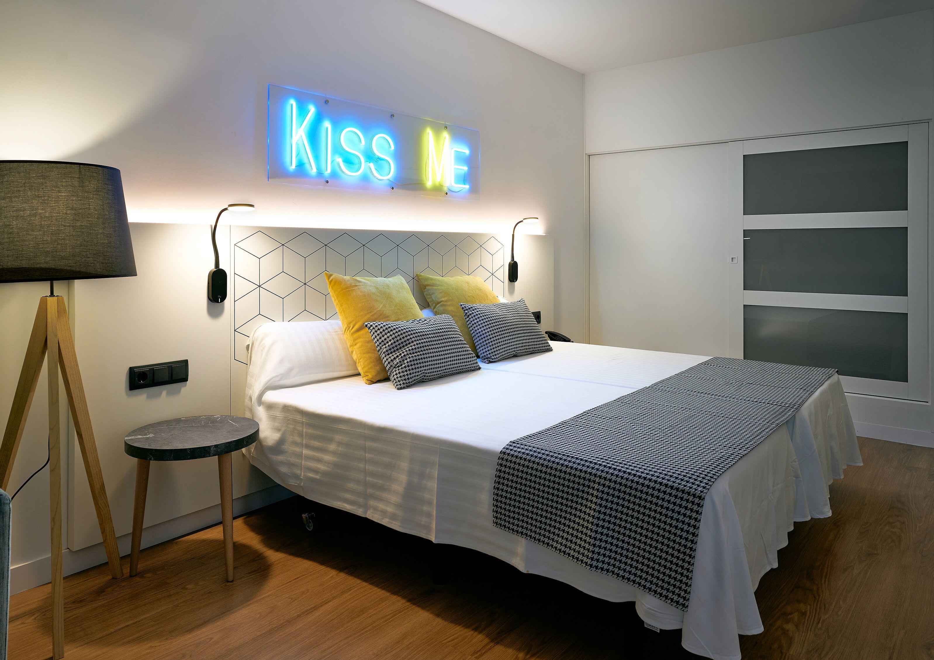 Un dormitorio moderno con cama, lámparas de noche y un letrero de neón que dice 'KISS ME'.
