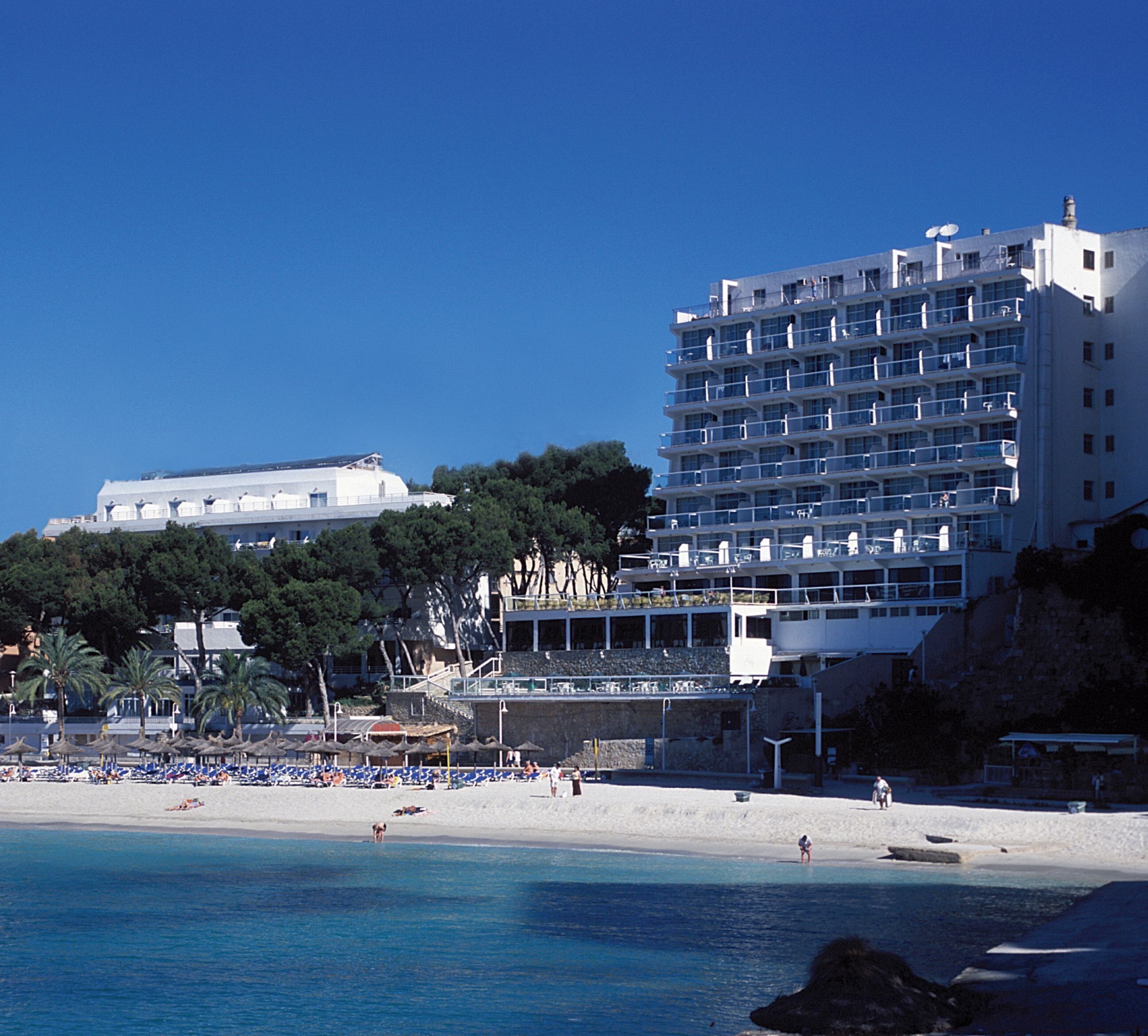 A coastal hotel with multiple balconies, a beach in the foreground, and a clear blue sky.