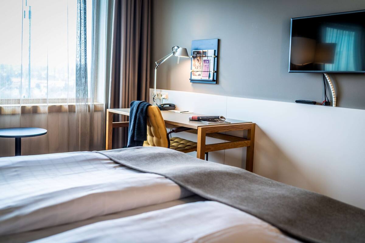 Quarto de hotel com cama, escrivaninha, cadeira, luminária, televisão e janela com cortinas.