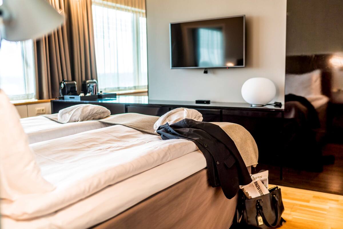 Quarto de hotel com cama, escrivaninha com TV de tela plana, luminária e janela com cortinas.