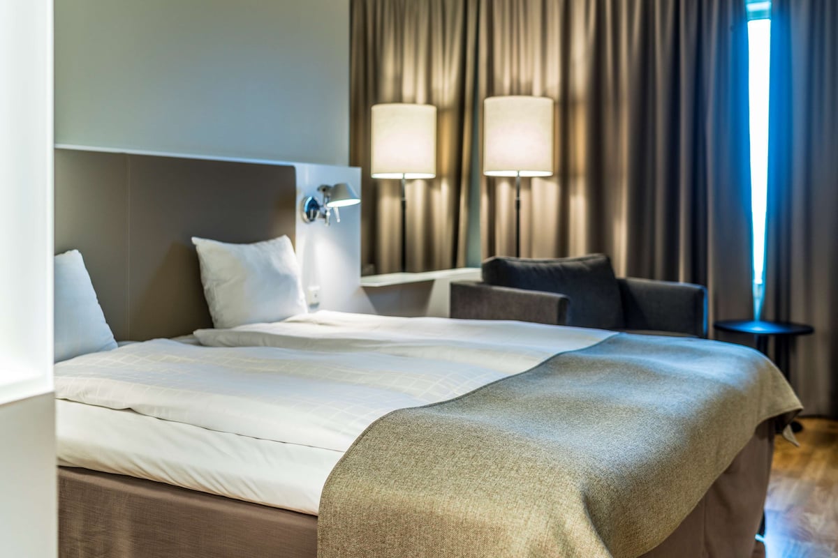 Quarto de hotel com uma cama grande, duas luminárias de cabeceira, uma cabeceira e vista para o exterior através das cortinas.