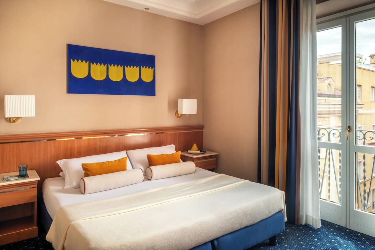 Habitación de hotel con cama, dos mesitas de noche, lámpara de pared, un cuadro y vistas a través de una puerta.