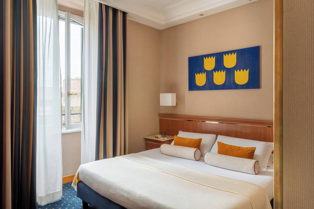 Una habitación de hotel con una cama grande, una mesita de noche, una lámpara de pared y un ventanal con cortinas.