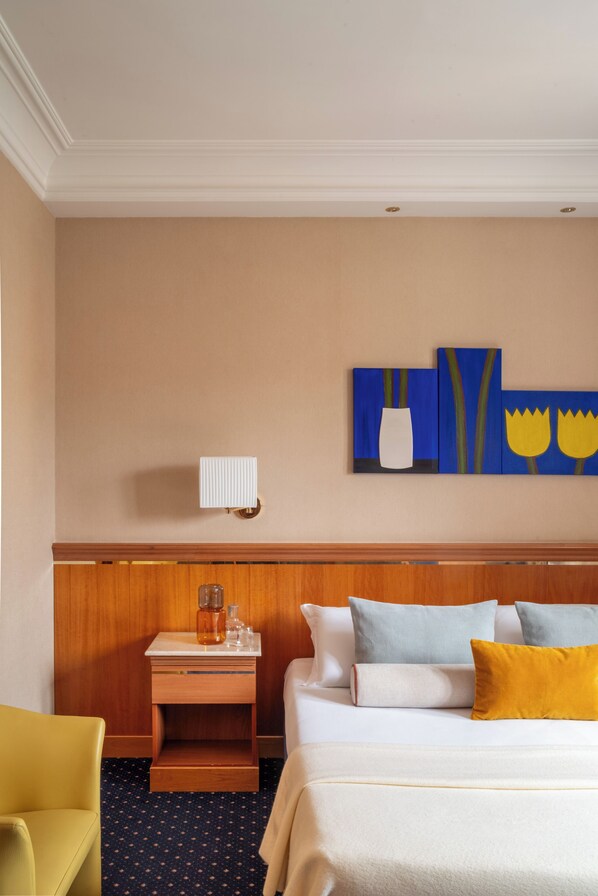 Una habitación de hotel con cama, mesita de noche, lámpara, una obra de arte en la pared y una silla.