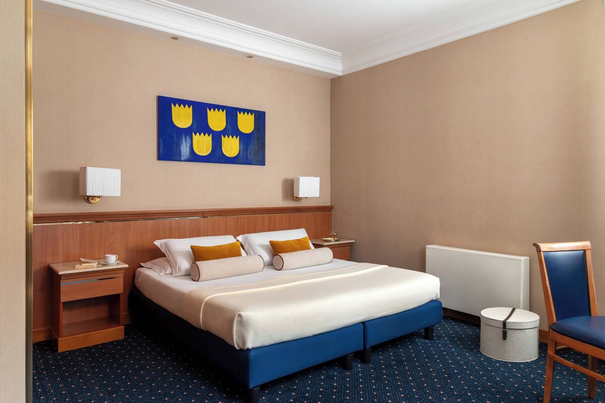 Una habitación de hotel con una cama, mesitas de noche, una silla, un radiador y una obra de arte en la pared.