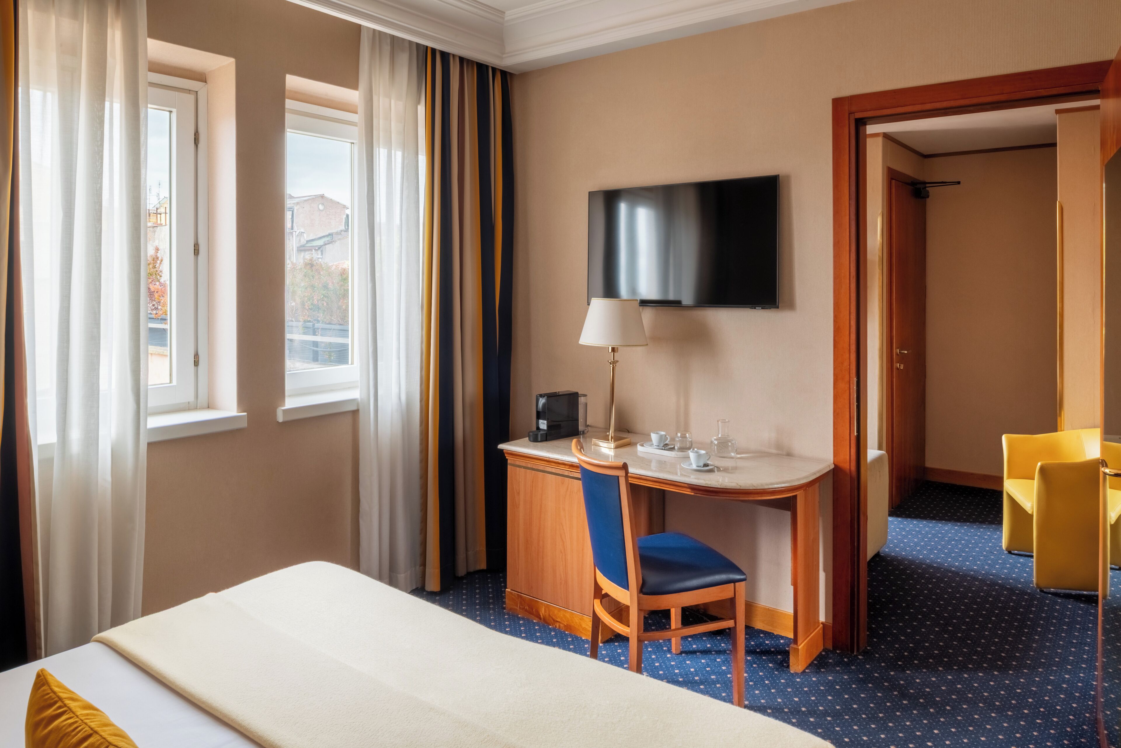 Habitació d'hotel amb un llit, una taula de treball amb una cadira, una televisió i una finestra amb cortines.