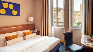 Ein Hotelzimmer mit Bett, Sessel, Schreibtisch, Fenster mit Vorhängen und einer Wand mit blauem Kunstwerk.