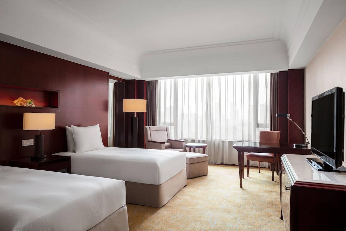 A hotel room with two beds, a desk, a TV, and a large window with curtains.