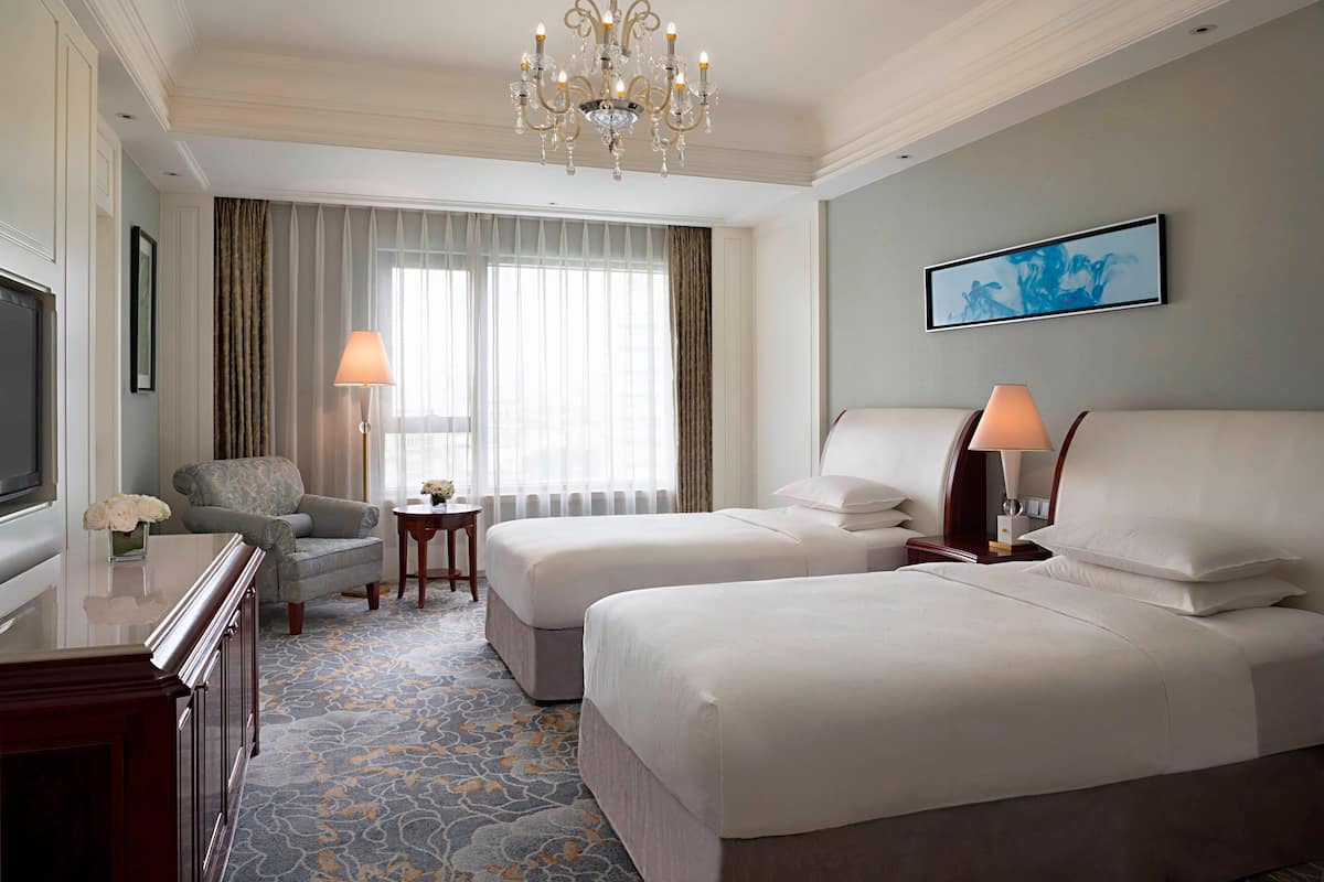 A hotel room with two beds, a TV, a desk, a chair, a chandelier, and a painting on the wall.