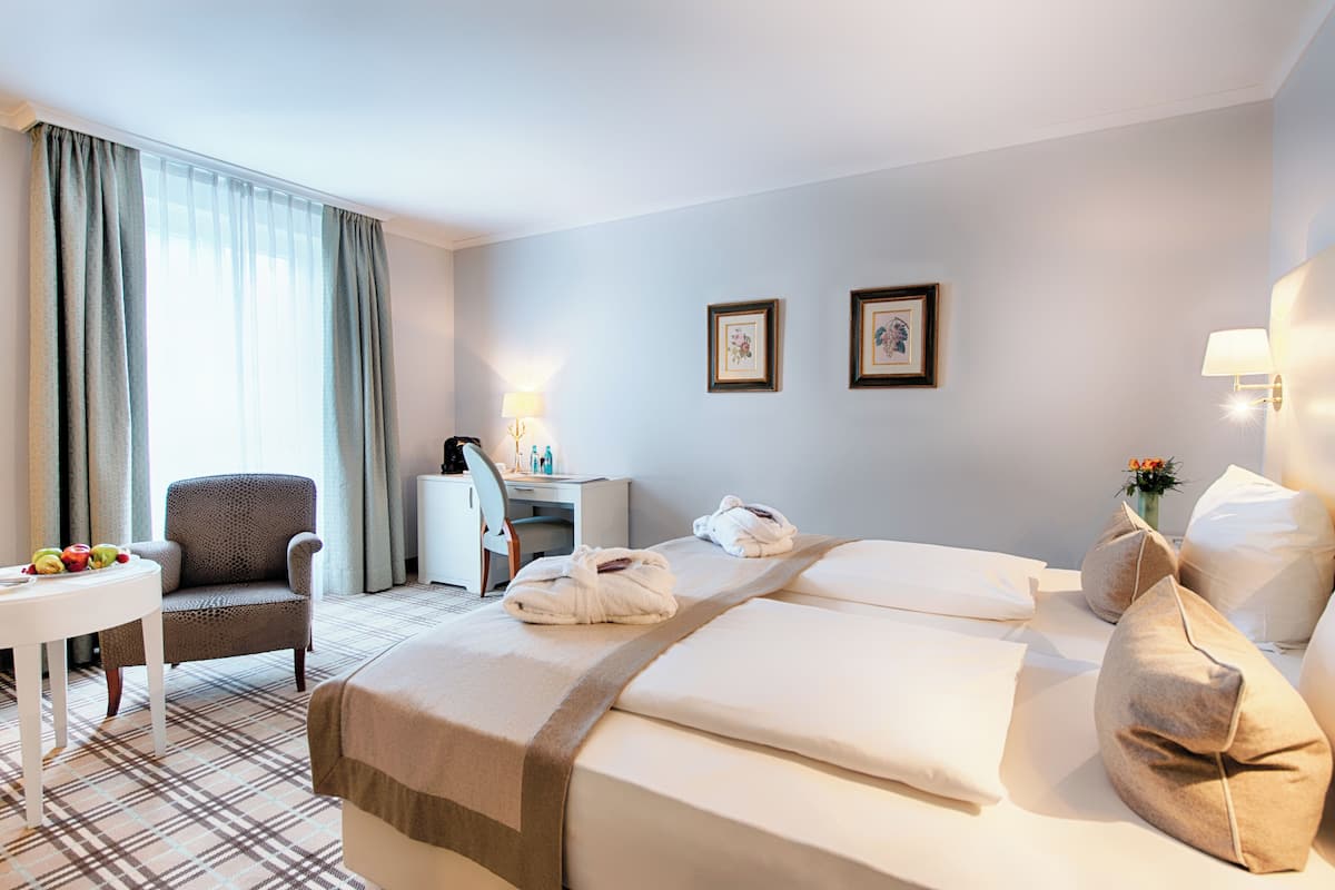 A hotel room with a bed, two chairs, a desk, a lamp, and a small table with fruit.