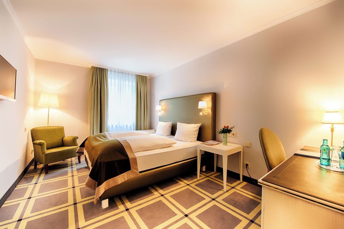 A hotel room with a bed, a desk, a chair, a lamp, and a window with curtains.