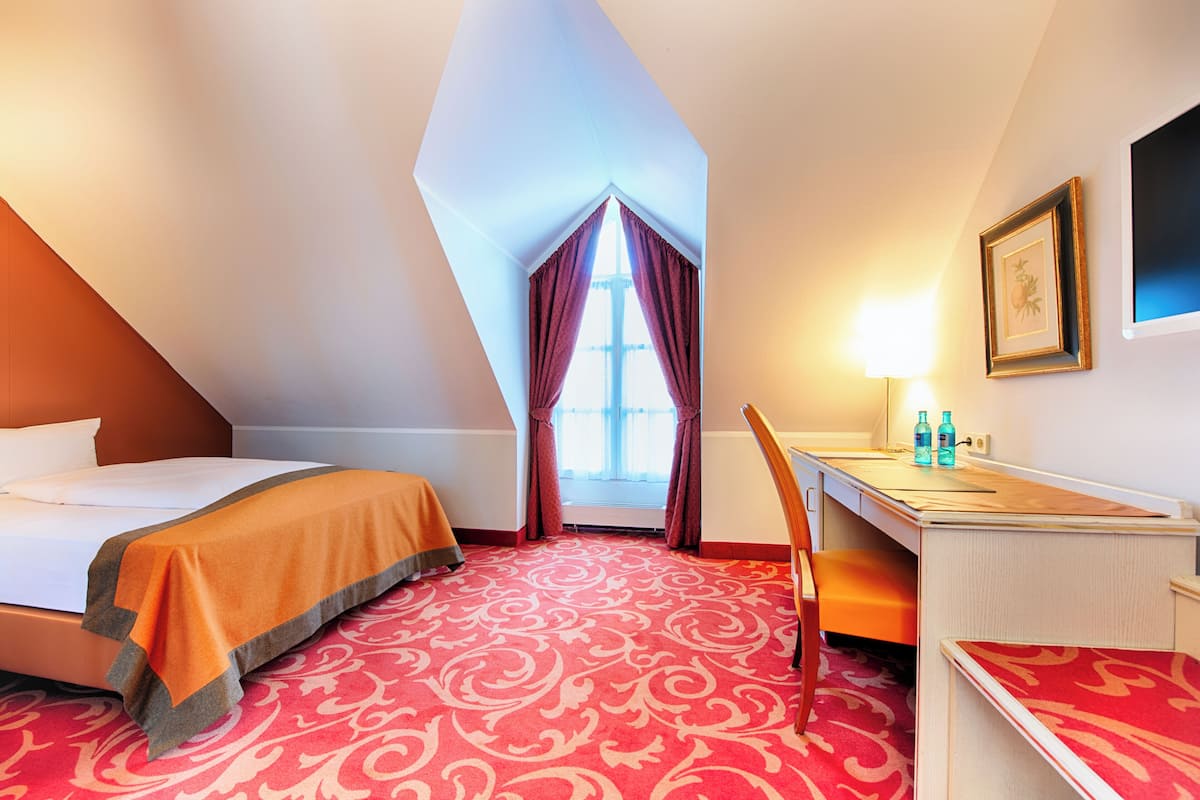 Une chambre d’hôtel avec un lit, un bureau, une chaise, une fenêtre avec des rideaux et une lampe.