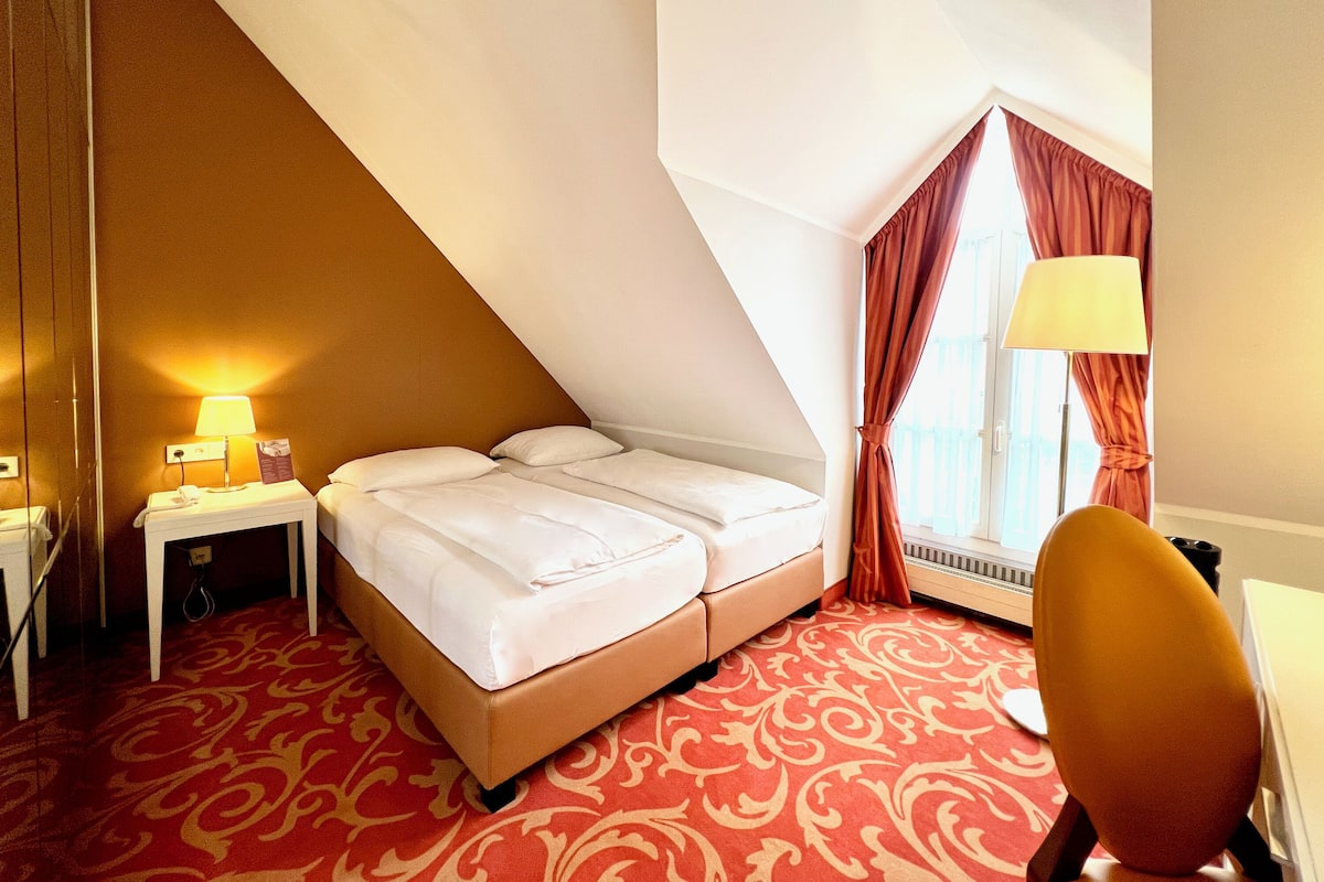 Une chambre d’hôtel avec un lit, un bureau, une chaise, une lampe et une fenêtre avec des rideaux.