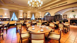 A spacious dining area with multiple round tables and chairs, a large chandelier, and a view of the outside through large windows.