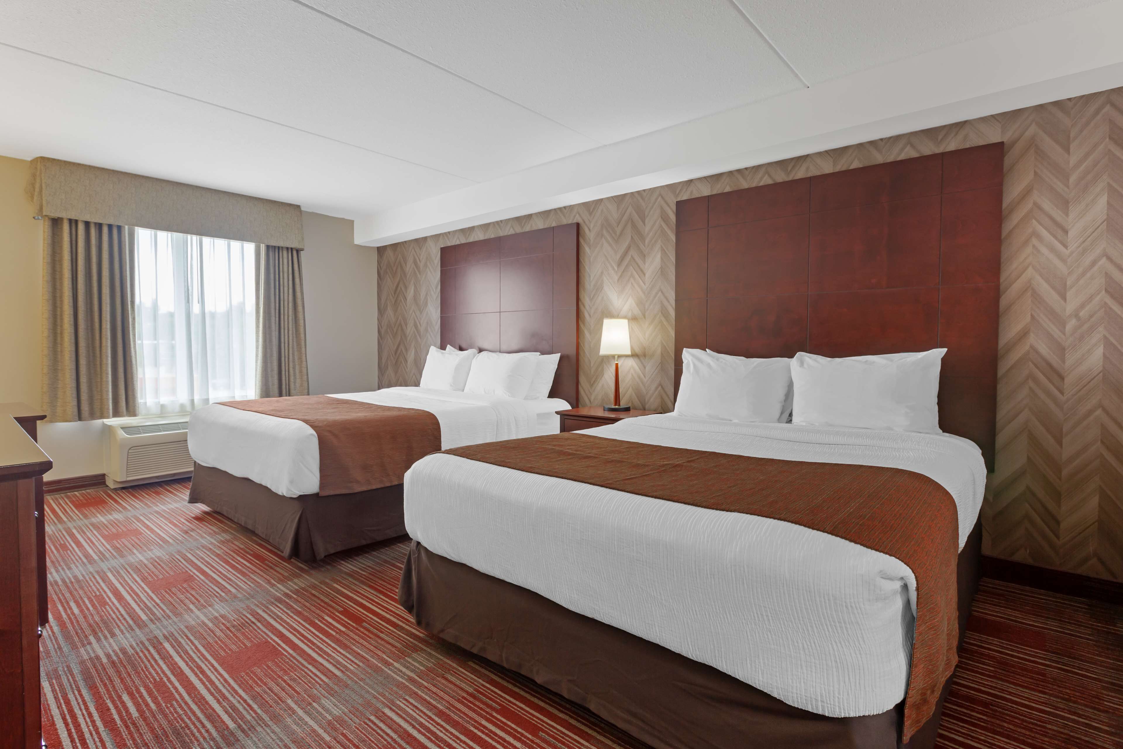 A hotel room with two beds, a desk, a lamp, and a large window with curtains.