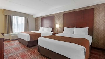 A hotel room with two beds, a desk, a lamp, and a large window with curtains.