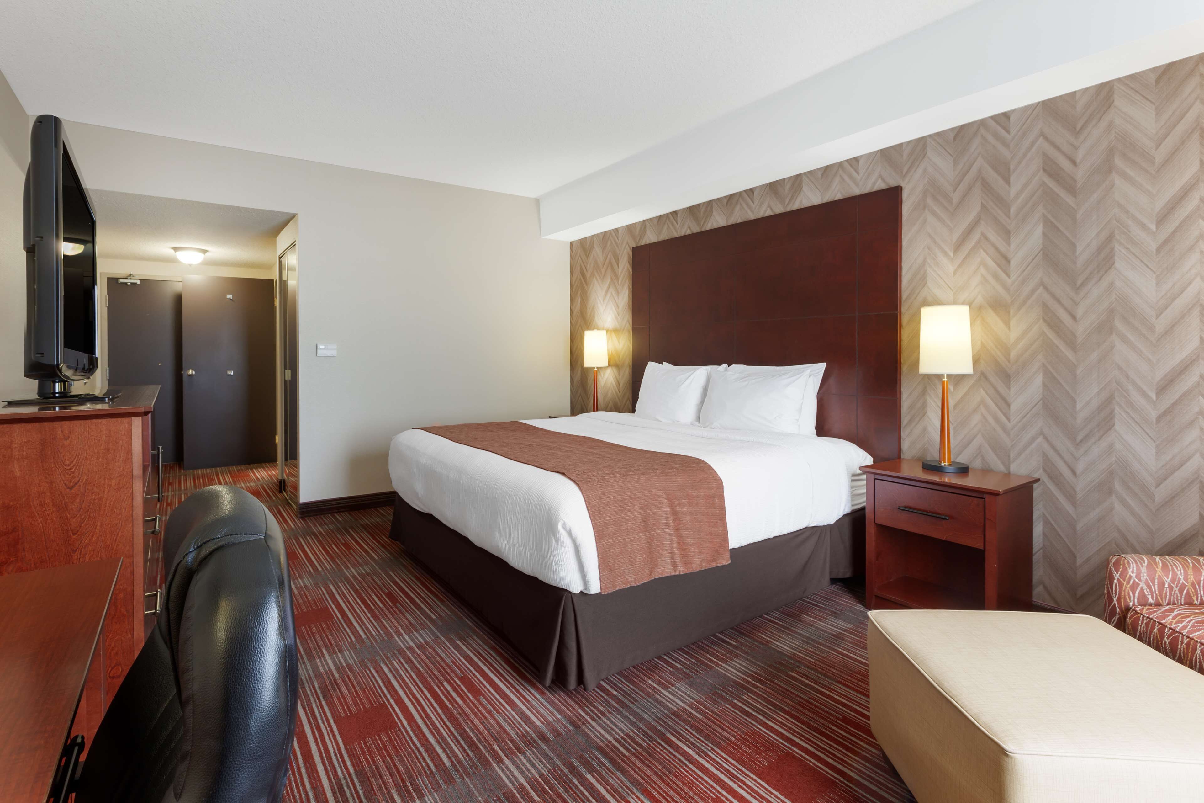 Hotelkamer met een groot bed, een bureau met een flatscreen televisie, een stoel, een nachtkastje met een lamp en een wand met een herringbone patroon.
