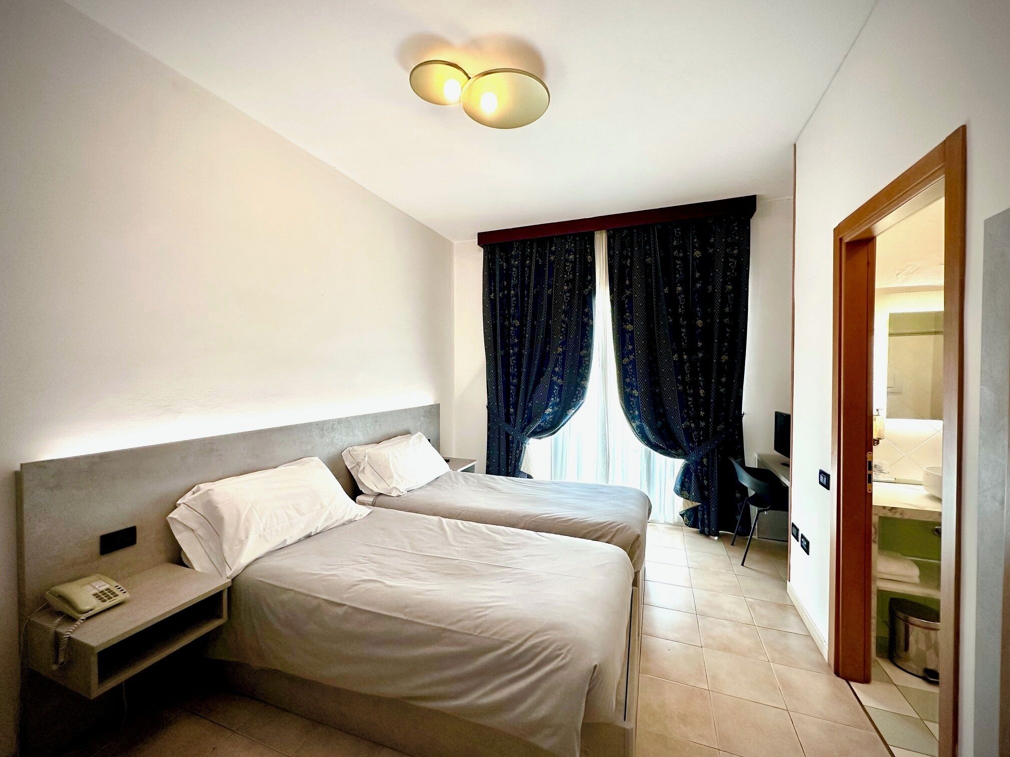 Ein Hotelzimmer mit zwei Betten, einem Nachttisch mit Telefon, einem Schreibtisch mit Stuhl und einem durchgehenden Türspalt sichtbaren Badezimmer.