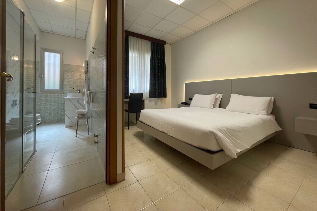 A modern hotel room with a large bed, a desk, and a bathroom visible through a glass door.