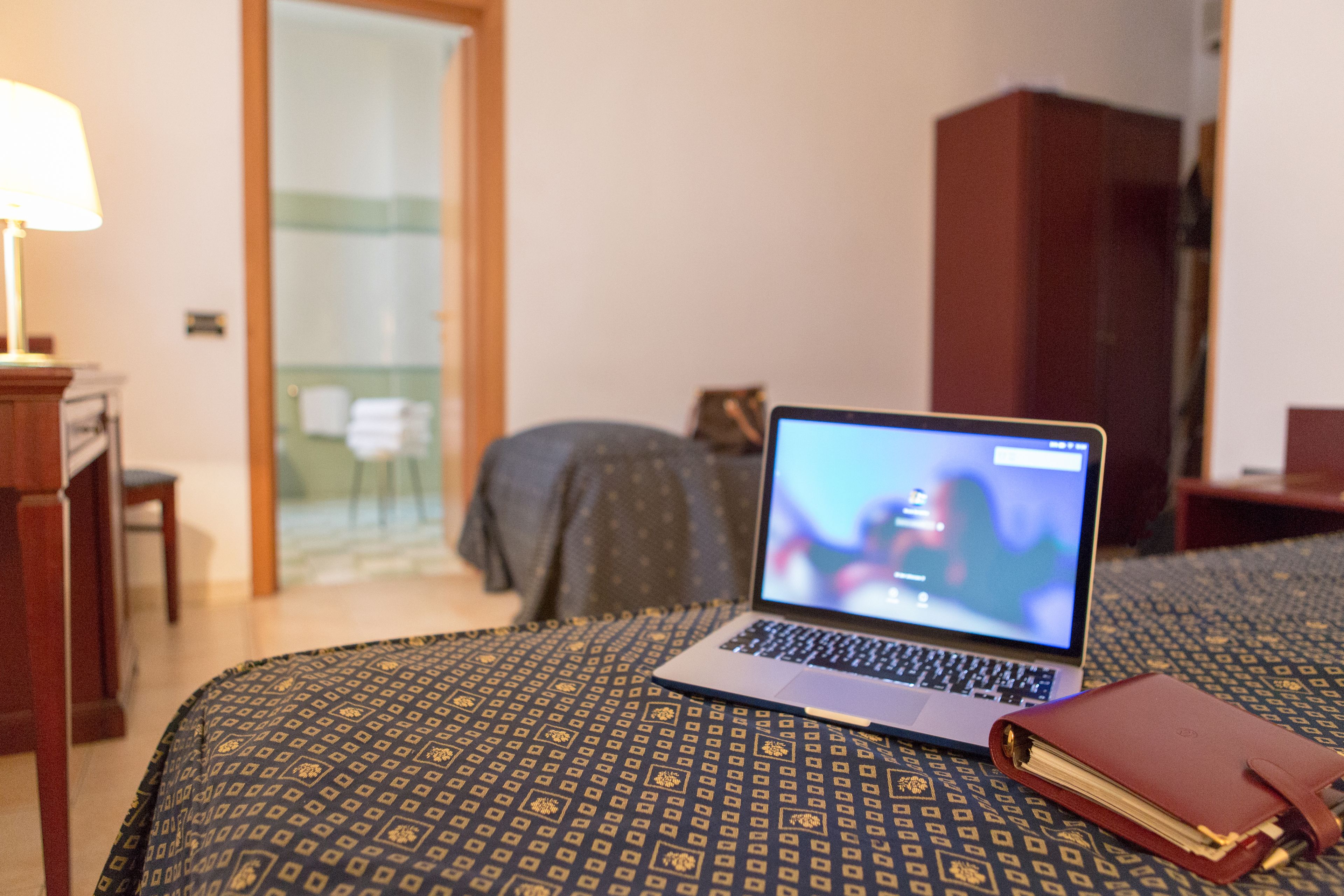 Un ordinateur portable est posé sur une couvre-lit à motifs dans une chambre d’hôtel, où l’on aperçoit également une table de chevet en bois, une lampe et une armoire.