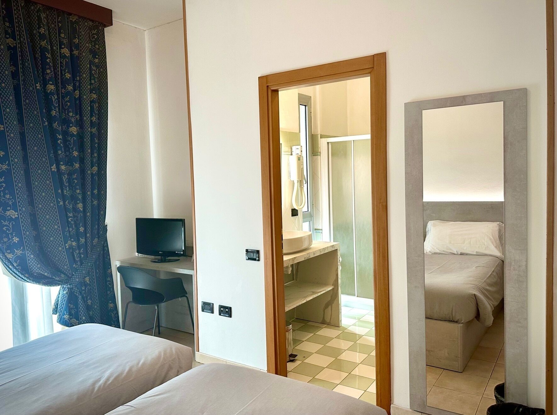 Ein Hotelzimmer mit zwei Betten, einem Schreibtisch mit Computer, einem Badezimmer mit Waschbecken und Spiegel sowie einer Dusche.