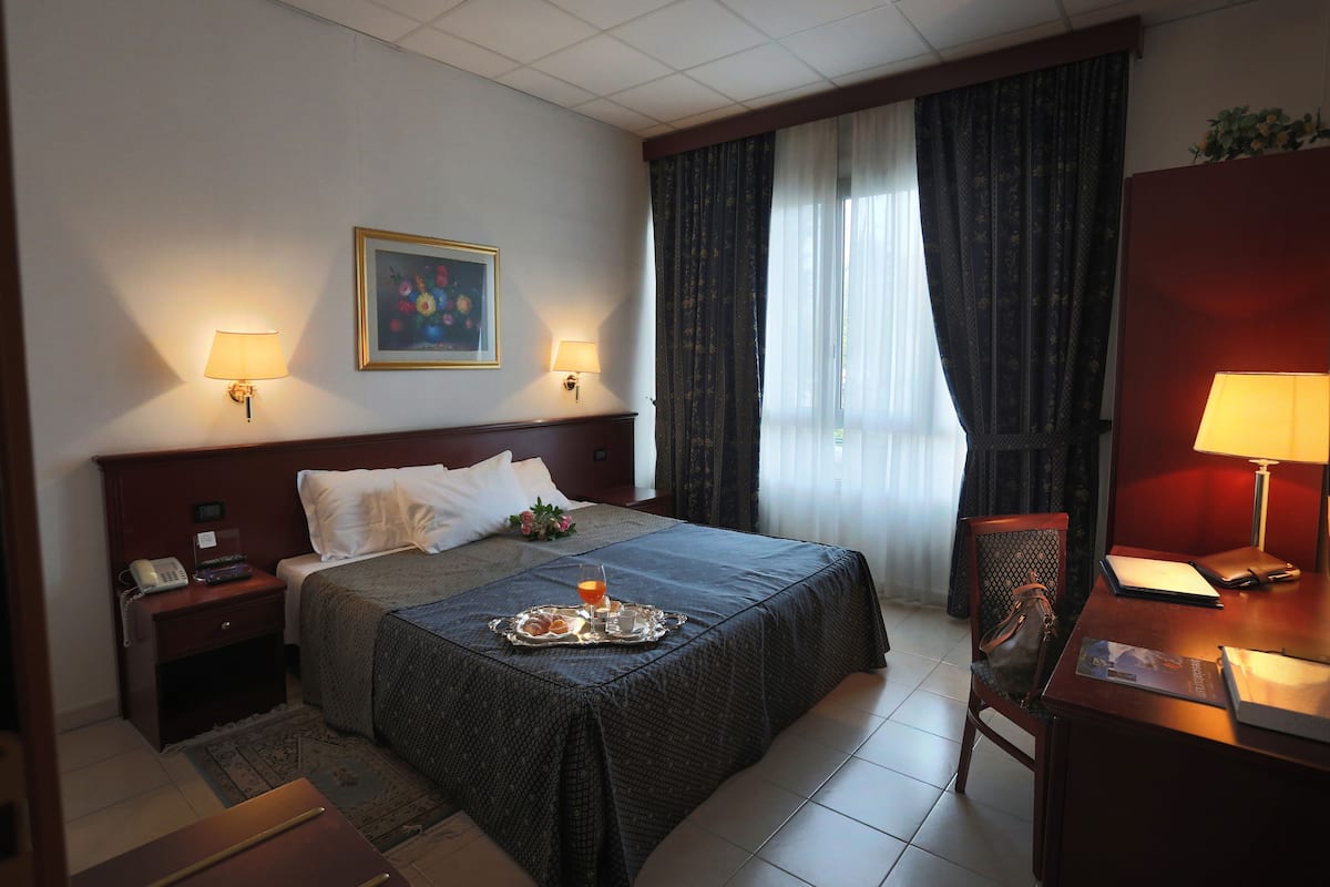 A hotel room with a bed, two bedside lamps, a desk with a chair, a small table with a tray of food and drink, and a painting on the wall.
