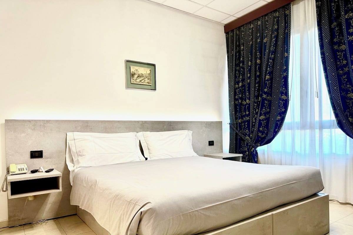 A modern hotel room with a large bed, a bedside table with a phone and remote controls, a framed picture on the wall, and a window with blue curtains.