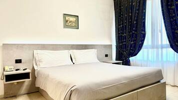 A modern hotel room with a large bed, a bedside table with a phone and remote controls, a framed picture on the wall, and a window with blue curtains.