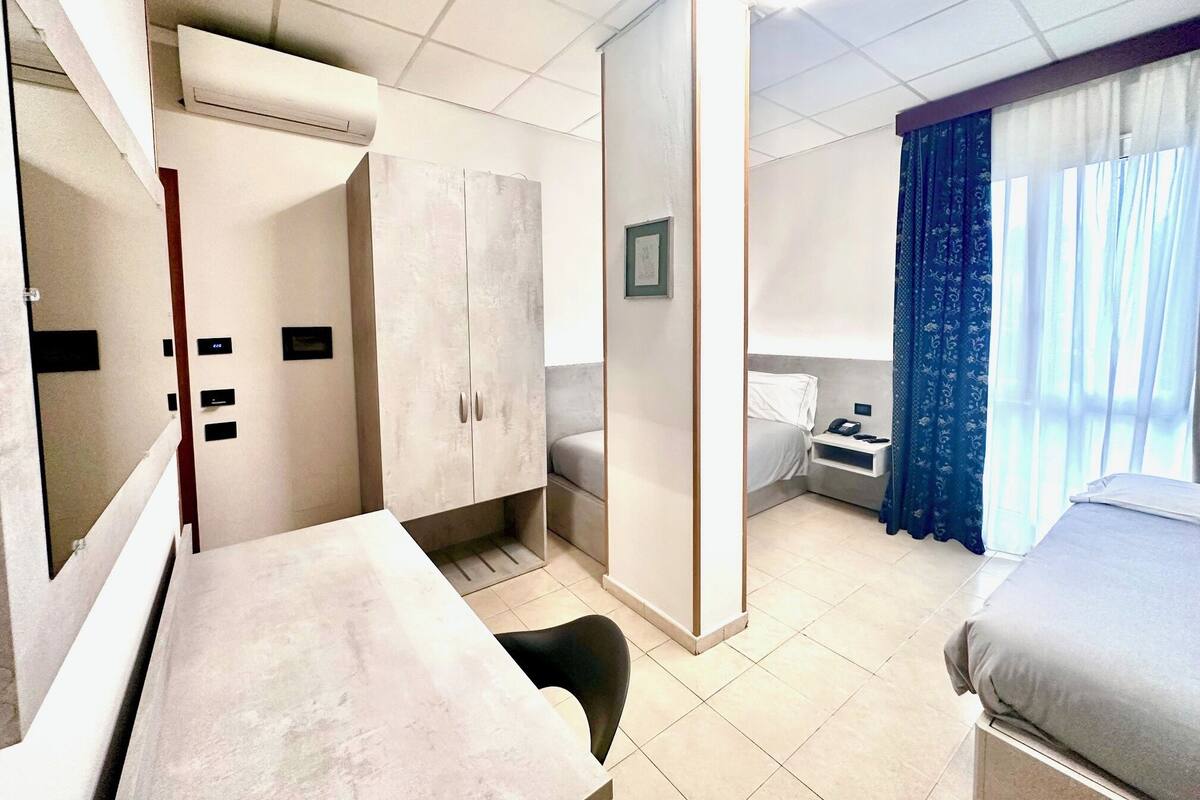 A hotel room with a bed, a desk, a wardrobe, a nightstand, and a window with curtains.