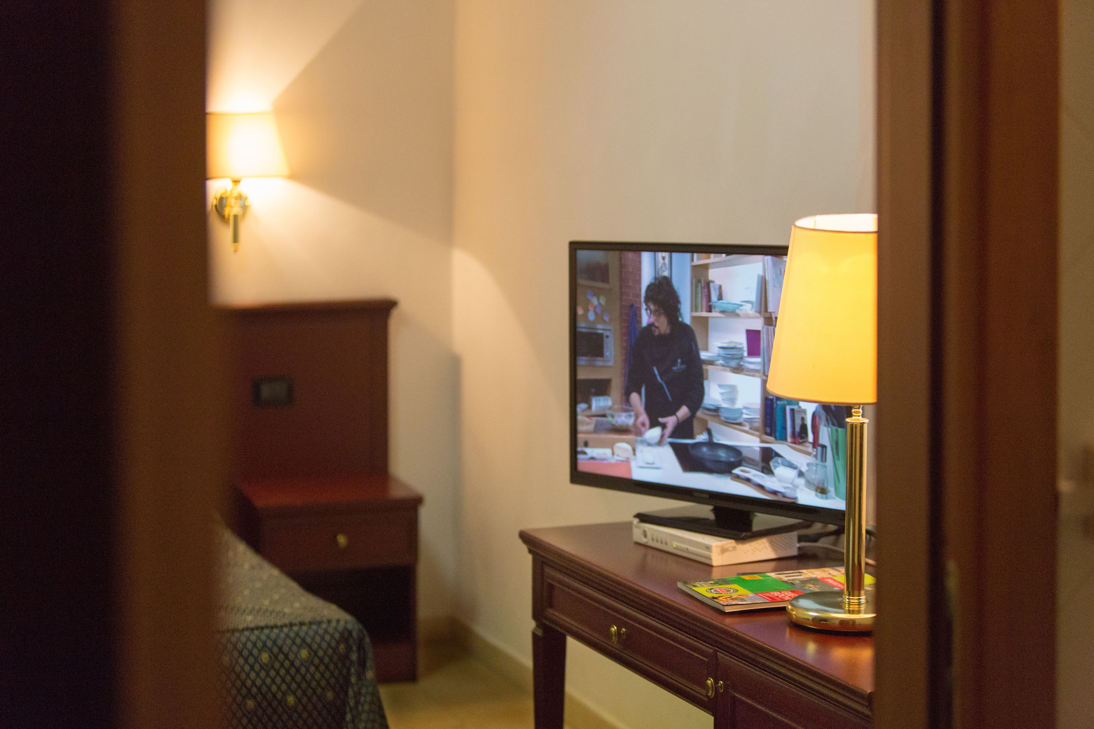 Quarto de hotel com televisão sobre uma mesa de madeira, uma luminária e uma pequena escrivaninha com gavetas.