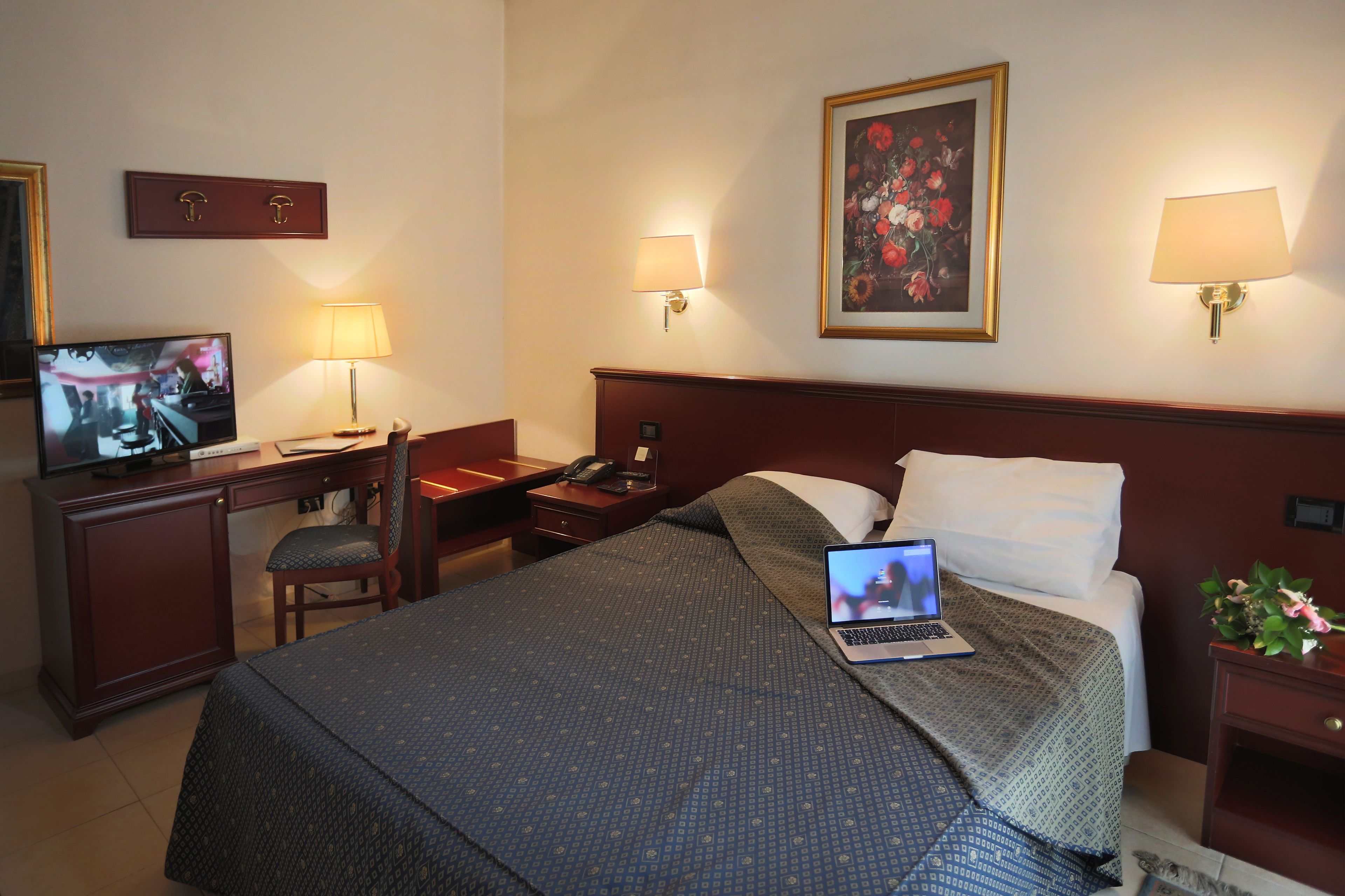 A hotel room with a bed, a desk with a laptop, a television, and a nightstand with a lamp.