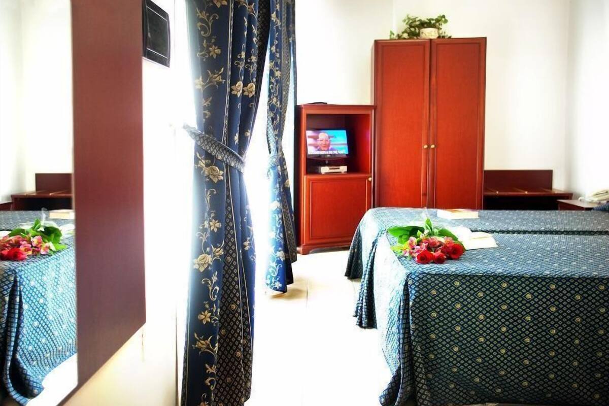 A hotel room with two beds, a wooden wardrobe, a television, and a window with curtains.