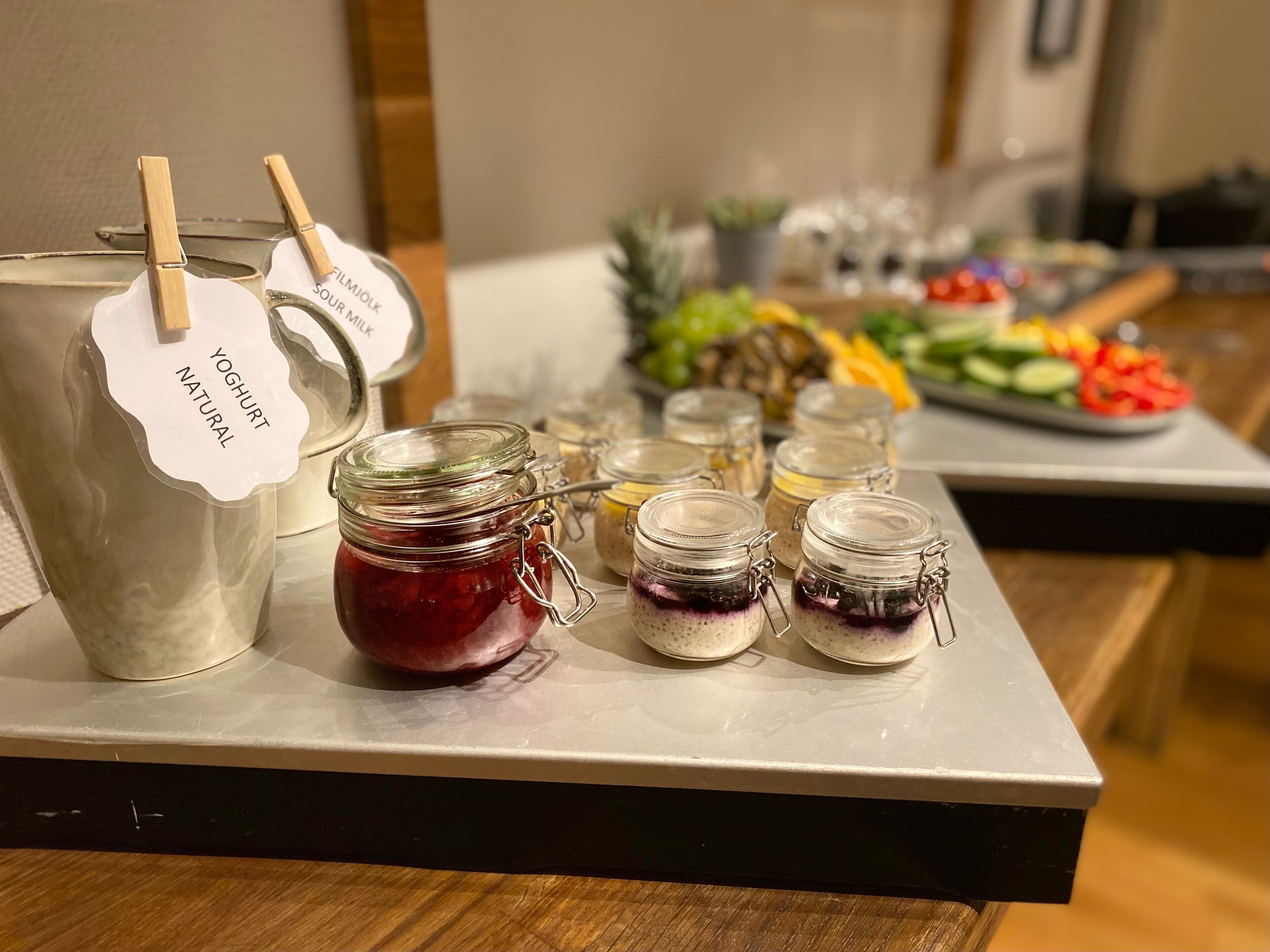 Una mesa de buffet con una jarra grande de yogur natural, varios frascos de mermeladas de frutas y una variedad de frutas y verduras frescas.