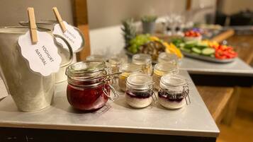 Una mesa de buffet con una jarra grande de yogur natural, varios frascos de mermeladas de frutas y una variedad de frutas y verduras frescas.