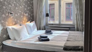 Un lit bien fait, avec du linge de lit blanc et gris, une tête de lit matelassée et une vue sur un bâtiment à travers une fenêtre ornée de rideaux.