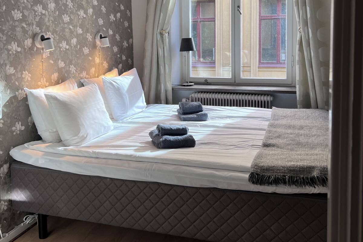 Cama bem arrumada com lençóis brancos e cinzas, cabeceira acolchoada e vista de um prédio através de uma janela com cortinas.