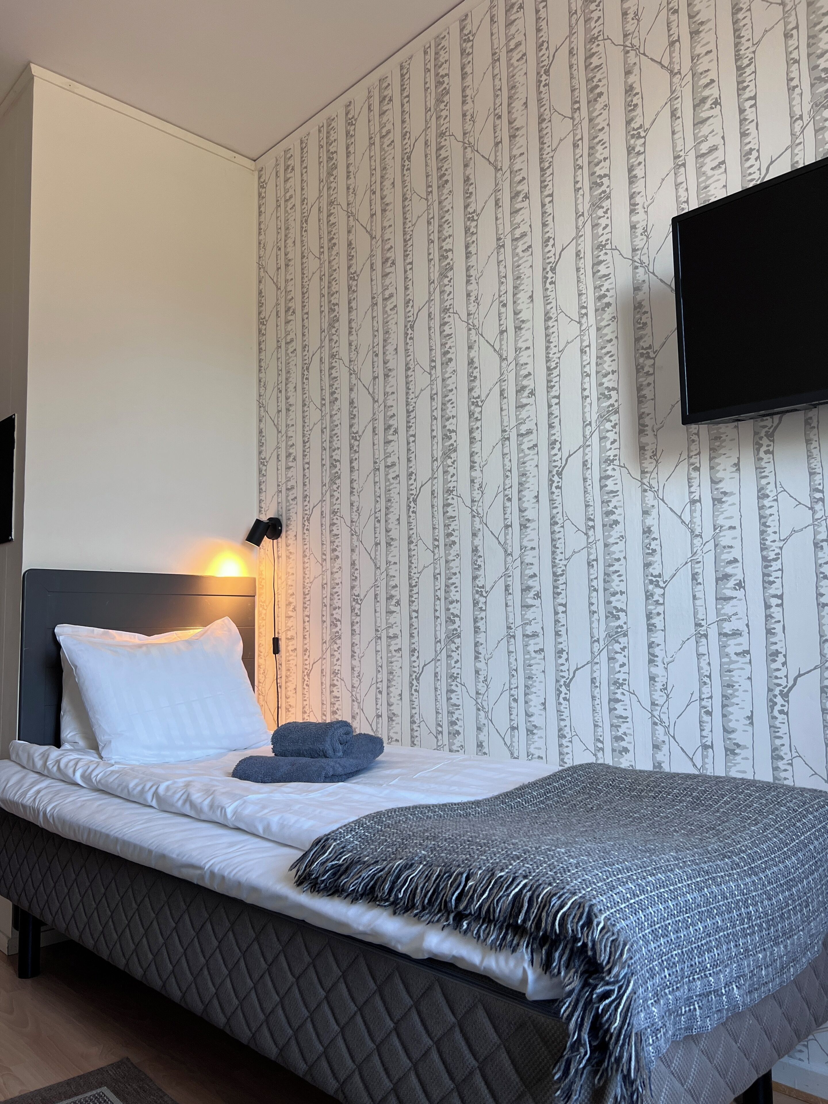 Una cama bien hecha con cabecera acolchada, un televisor montado en la pared y un papel tapiz estampado.