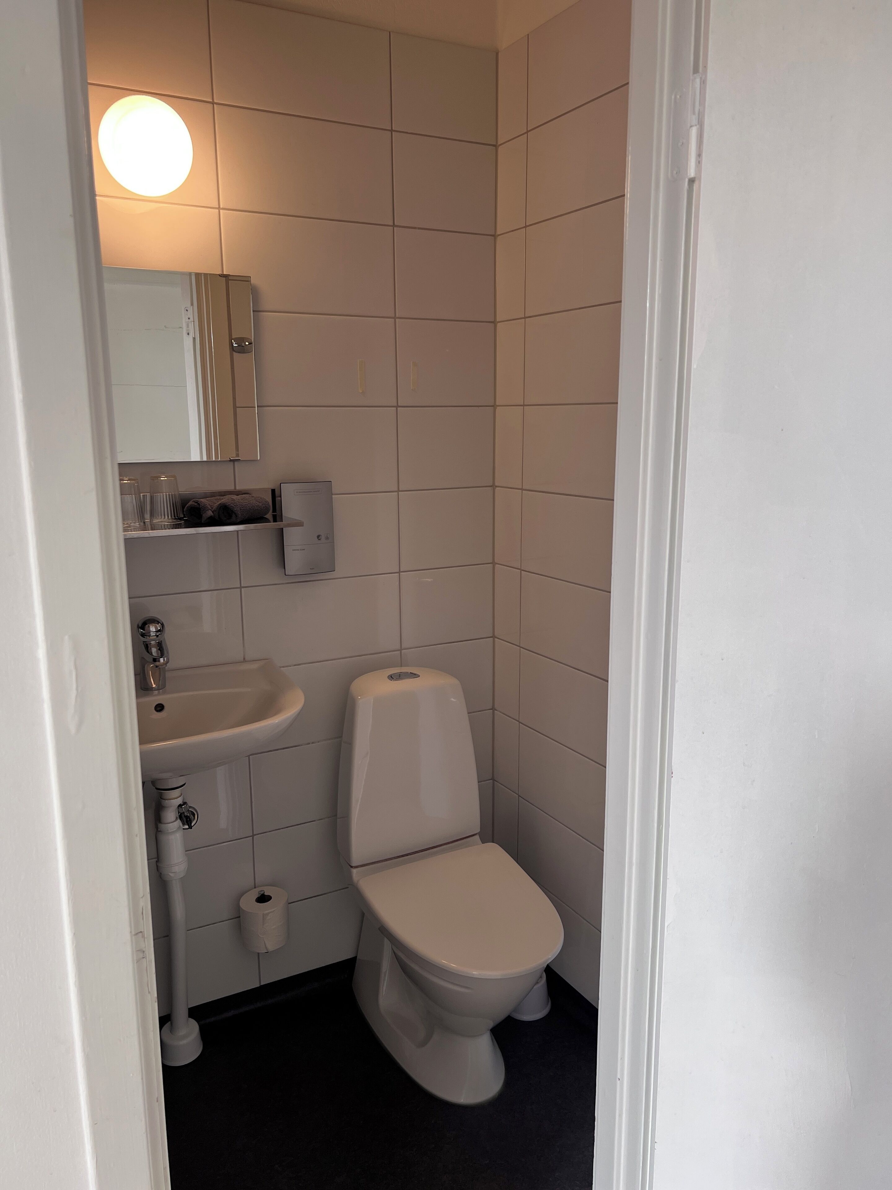 Un baño pequeño y limpio con azulejos blancos, un lavamanos, un inodoro y un espejo.