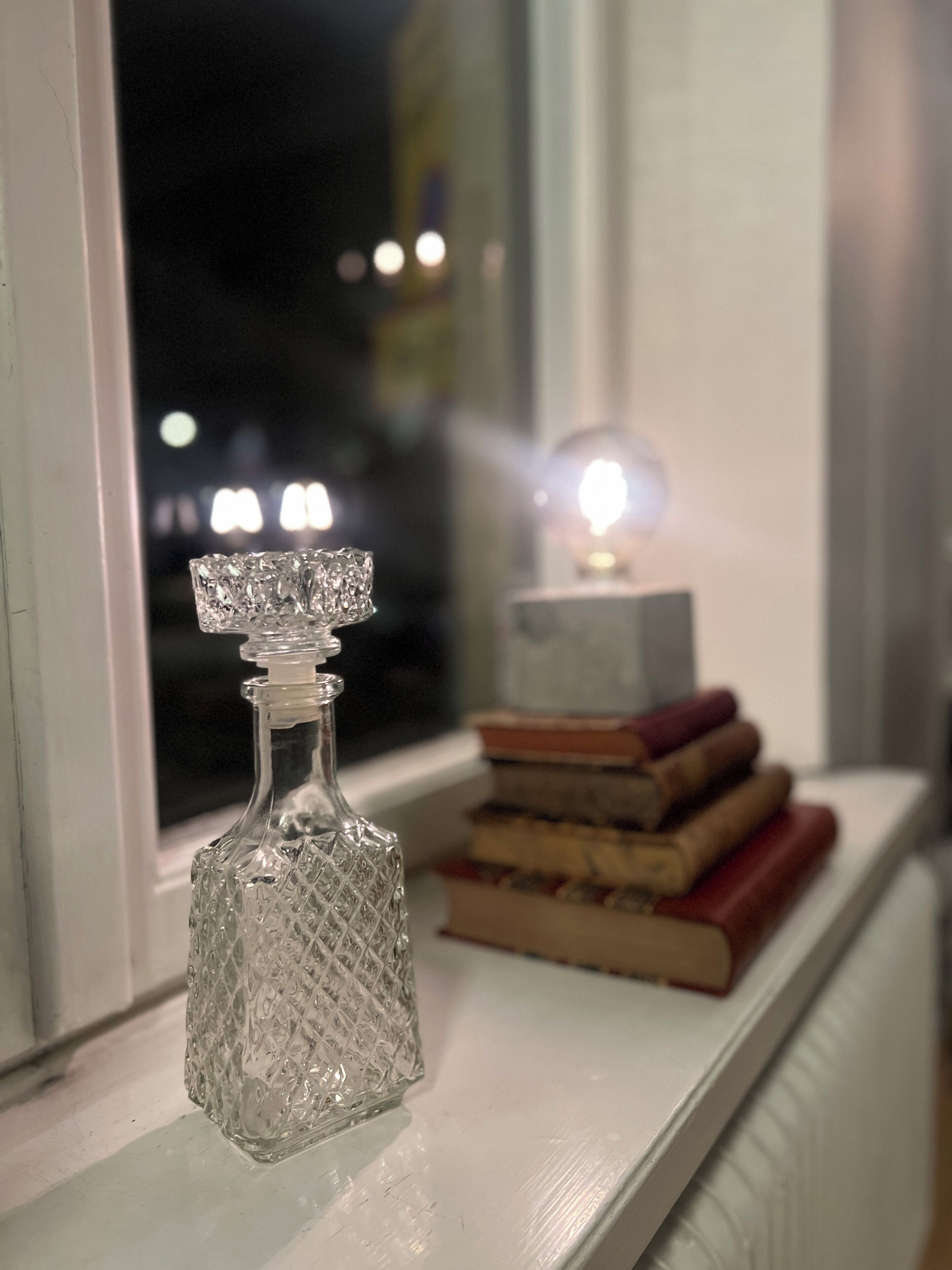 Une bouteille en verre transparent, ornée d’un motif en relief, est posée sur un rebord de fenêtre, à côté d’une pile de livres et d’une petite lampe.