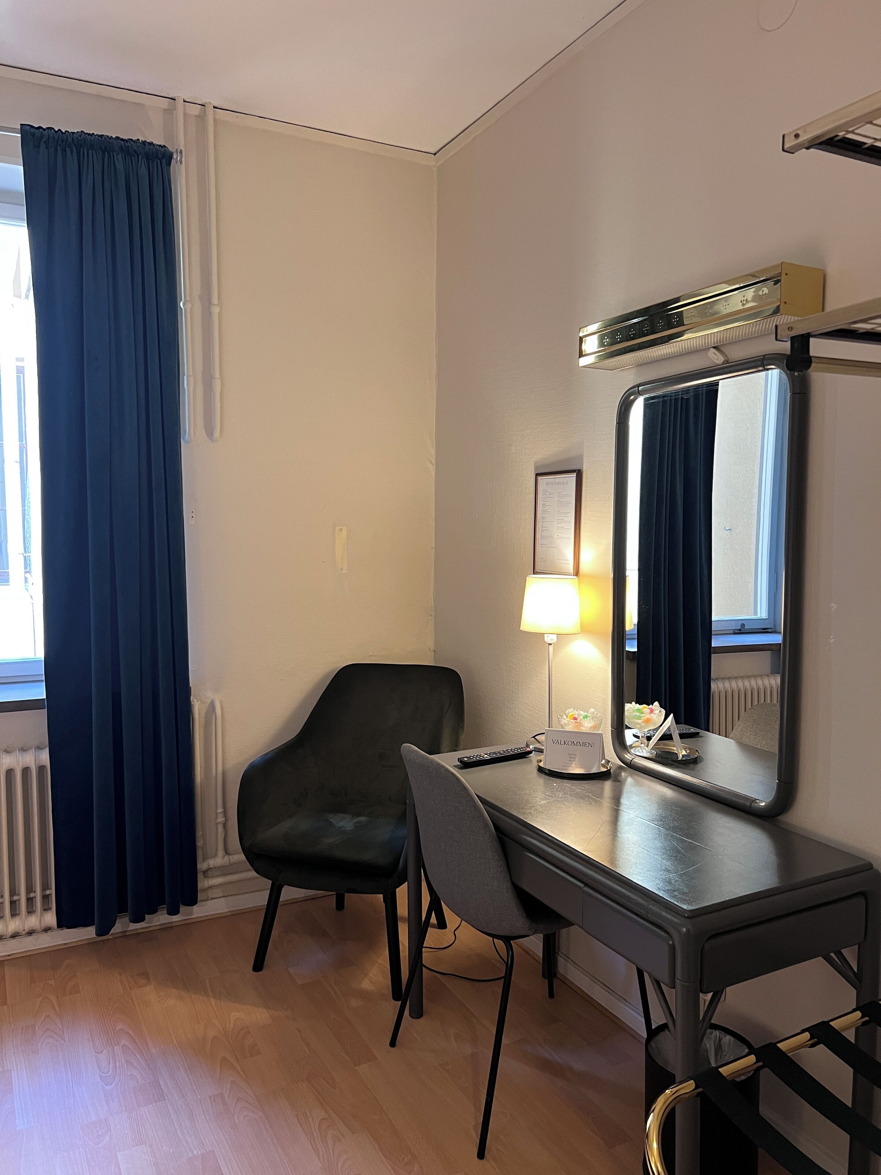 A hotel room with a desk, two chairs, a mirror, a lamp, and a radiator.