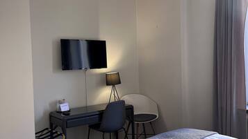 Quarto de hotel com cama, escrivaninha com cadeira, televisão na parede e janela com cortinas.