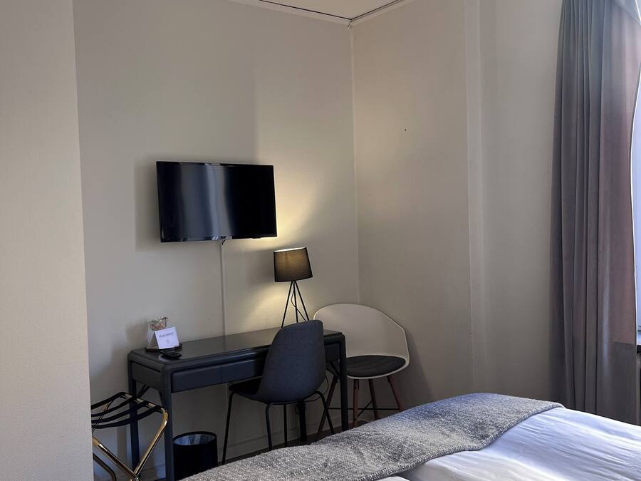 Habitación de hotel con cama, escritorio con silla, televisor montado en la pared y ventana con cortinas.