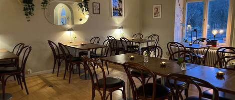 A dining area with wooden tables and chairs, a large mirror, and various wall decorations including framed pictures and plants.