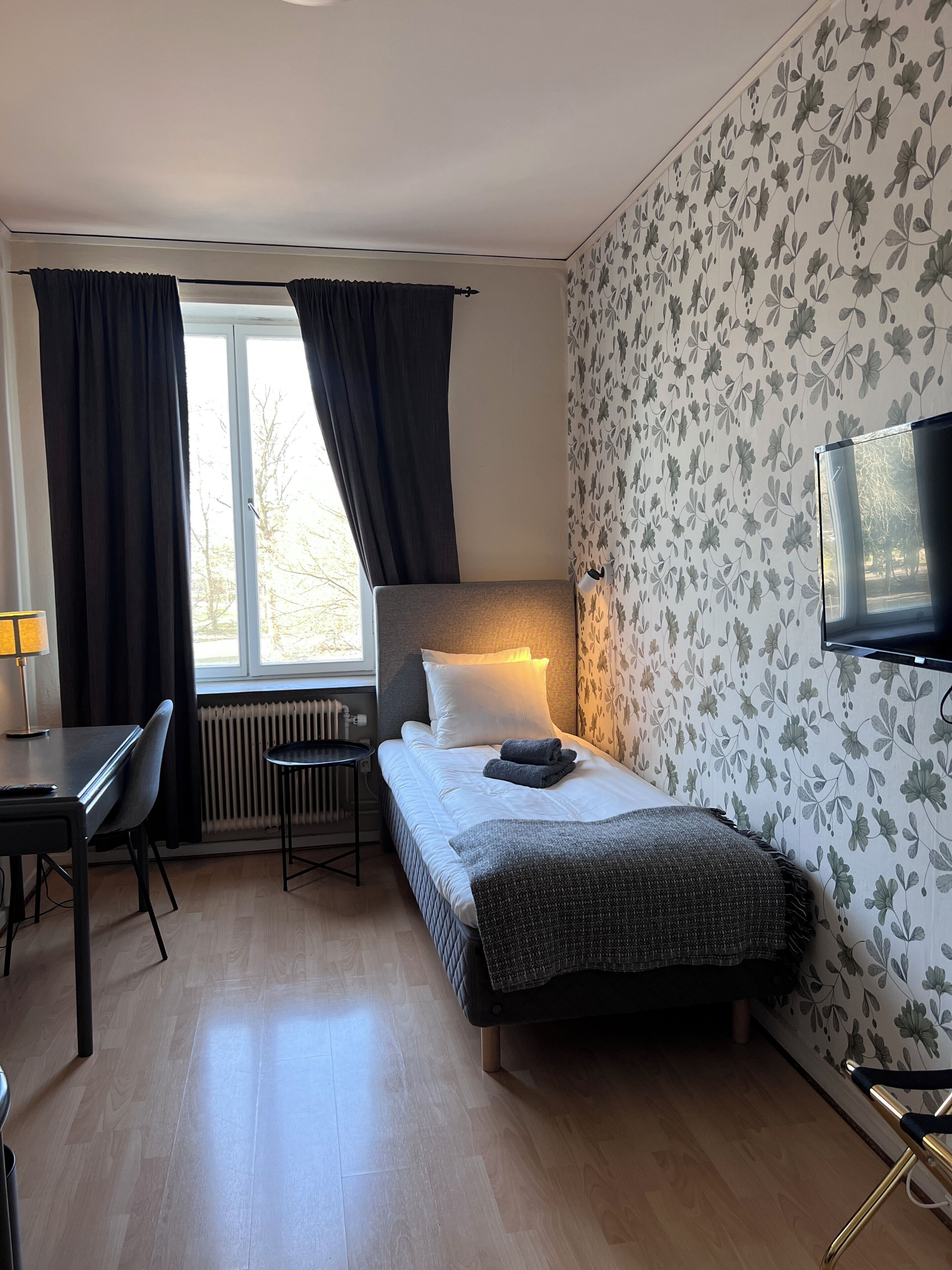 Habitación de hotel con cama, escritorio, silla, televisor y ventana con cortinas.
