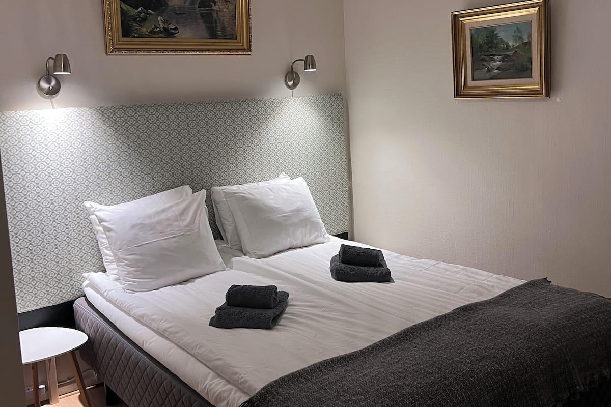 Uma cama bem arrumada com lençóis brancos e cabeceira acolchoada escura, duas luminárias fixadas na parede e dois quadros emoldurados nas paredes.