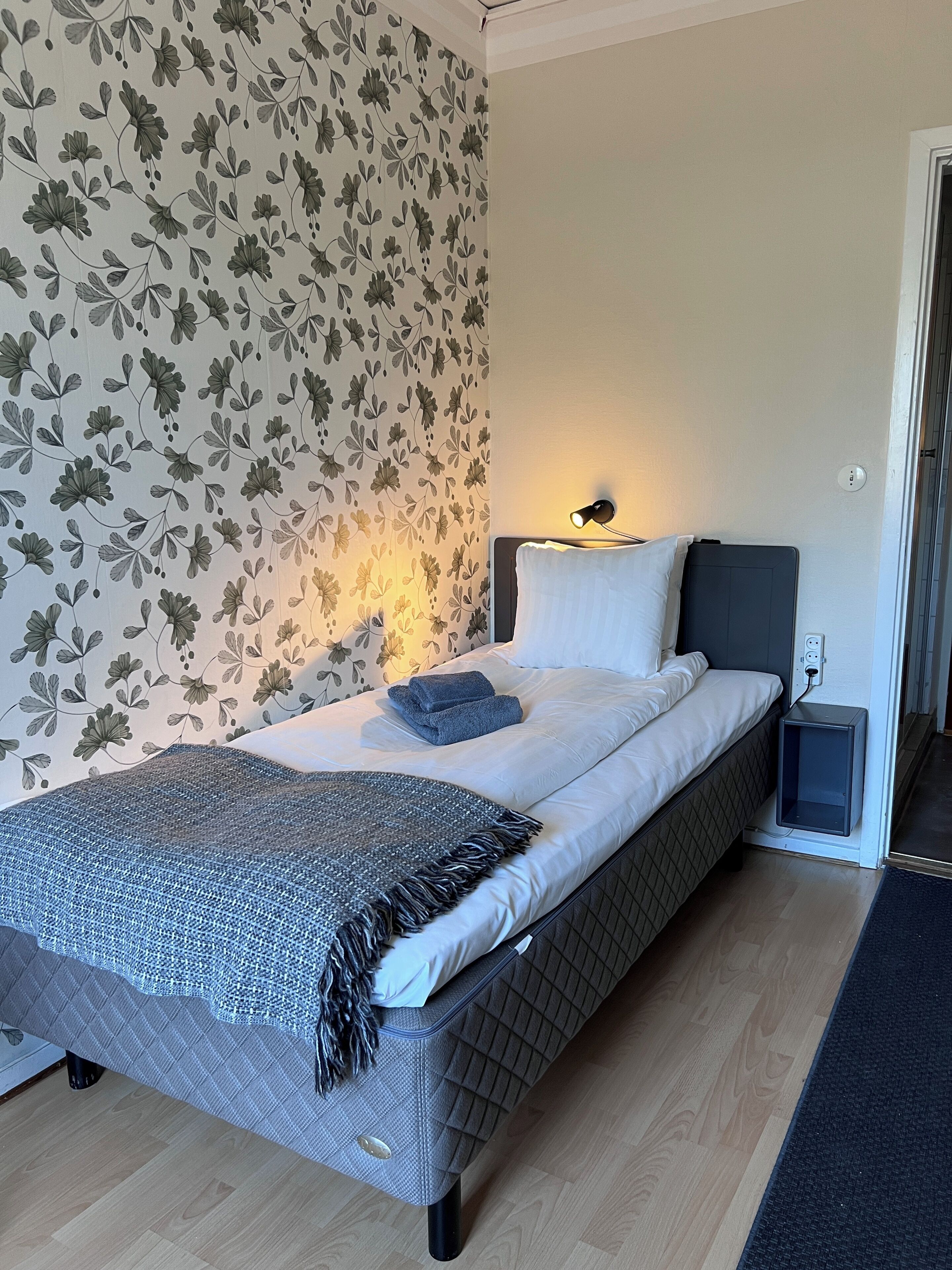 A neatly made bed with a grey headboard, a bedside lamp, and a floral wallpaper pattern.