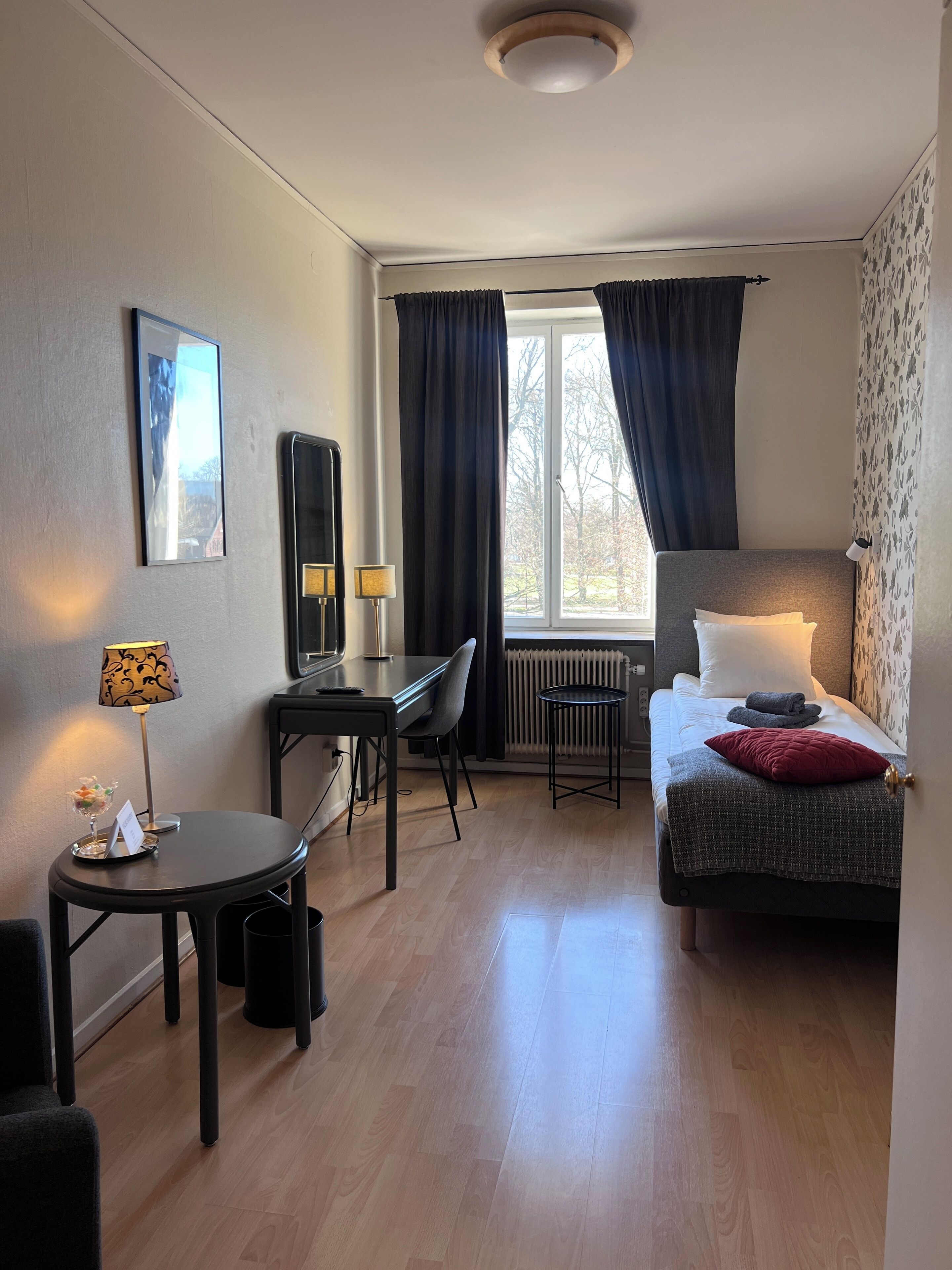 A compact hotel room with a bed, a desk, a chair, a small table, a lamp, and a radiator.